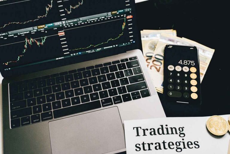 A laptop displaying trading charts, calculator, and money with "Trading Strategies" paper. Ideal for beginners learning the basics of trading and strategies.