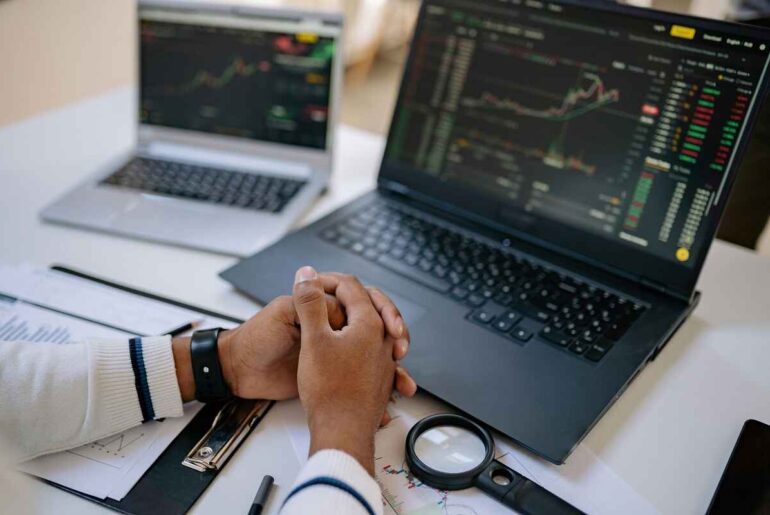 A person analyzing stock charts on dual laptop screens, surrounded by reports and tools. Ideal for understanding stock market analysis.