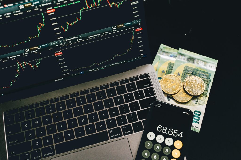 A laptop displaying stock charts, accompanied by cryptocurrency coins, euro bills, and a smartphone calculator, illustrating the concept of trading for penny stocks with real-time analysis and financial tools.