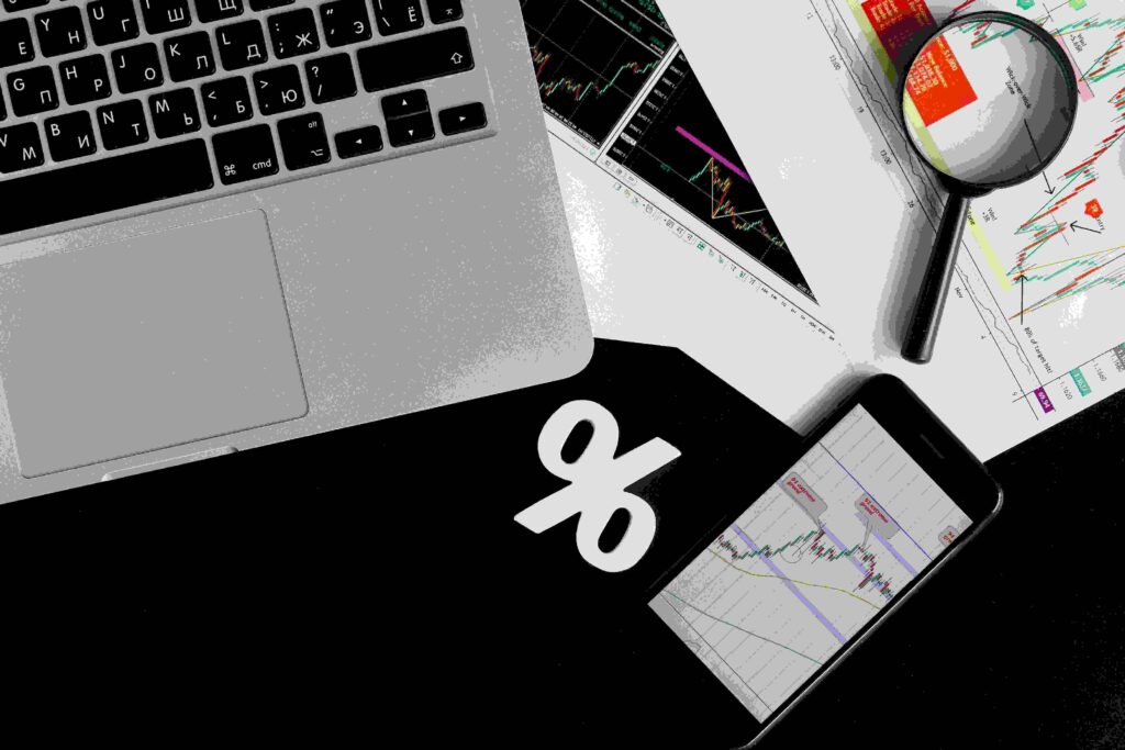 Image showing trading charts on a smartphone and papers, a laptop, magnifying glass, and a percentage sign, symbolizing trading analysis and risk management.