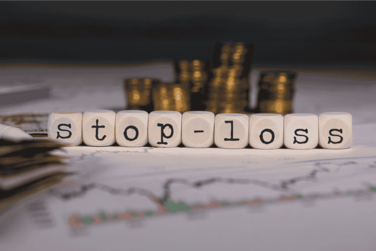 Stop-loss spelled out in wooden letter cubes on a stock market chart, with stacked gold coins in the background representing financial risk management and trading.